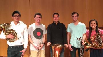 Five intercollegiate band members posing with musical instruments on stage.