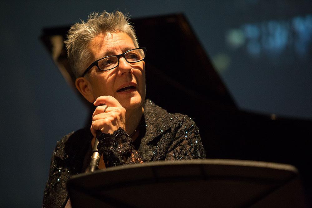 Person holding microphone, wearing glasses, with a piano in the background.