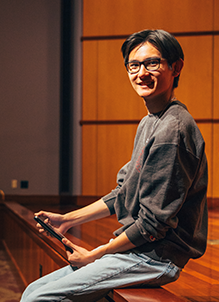 Male student sitting on the edge of a stage with a grand piano in the background