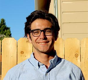 A person wearing glasses stands outside in front of a wooden fence.