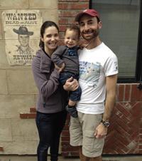 A smiling family stands outside a building with a 'Wanted' poster.