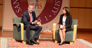 Bill de Blasio and another person seated, speaking at an event with a red backdrop. image link to story