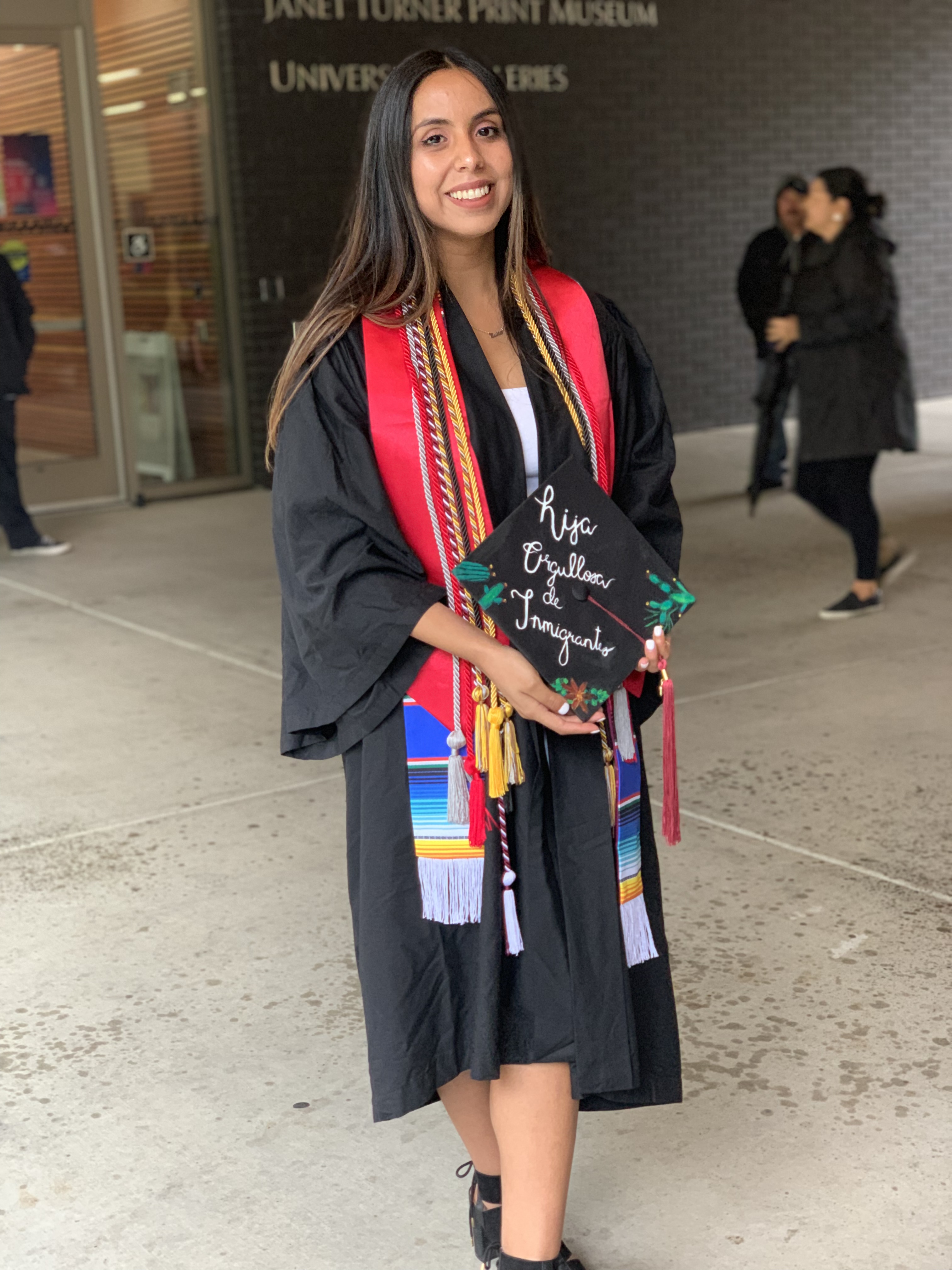 Young woman in graduation attire holding a diploma, standing outside a building. 