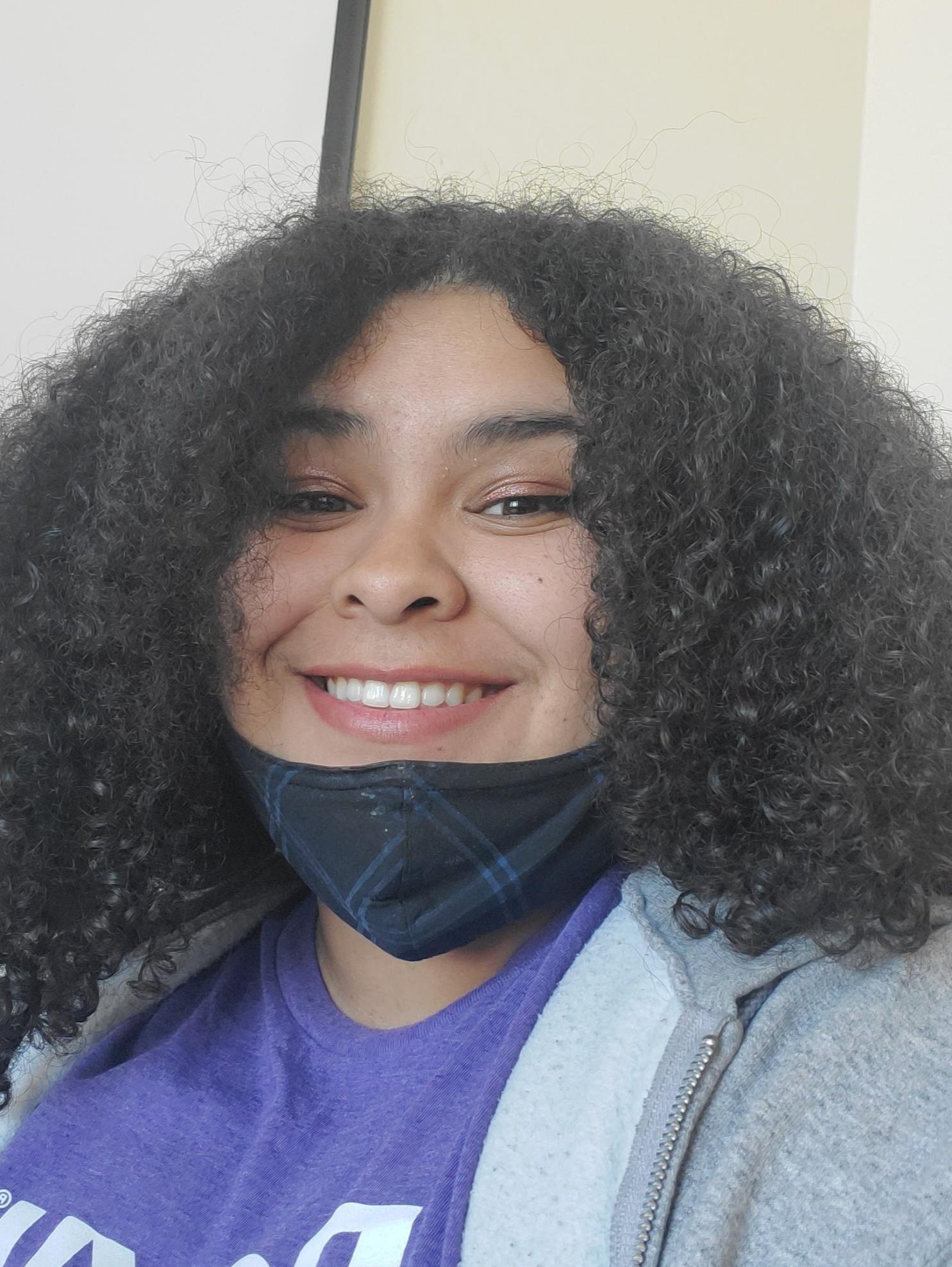 A person smiling, wearing a purple shirt and a gray jacket. 