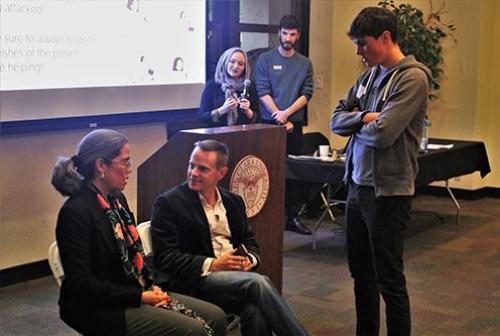 Two people sitting, one standing, with two people in background speaking at podium. image link to story