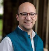A smiling person wearing glasses and a blue vest.