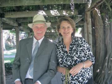 Two people sitting outdoors, one in a suit and hat, smiling.
