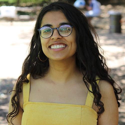 A person smiling outdoors, wearing glasses and a yellow top.
