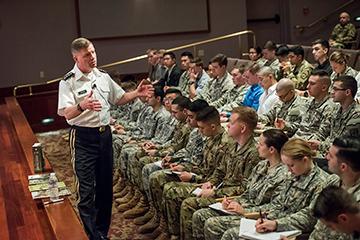 A military officer speaking to a seated group of soldiers.
