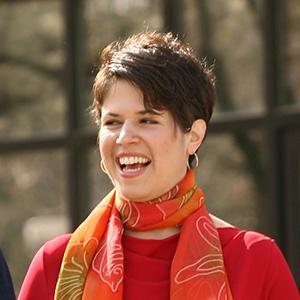 Person smiling and wearing red top and orange scarf outdoors.