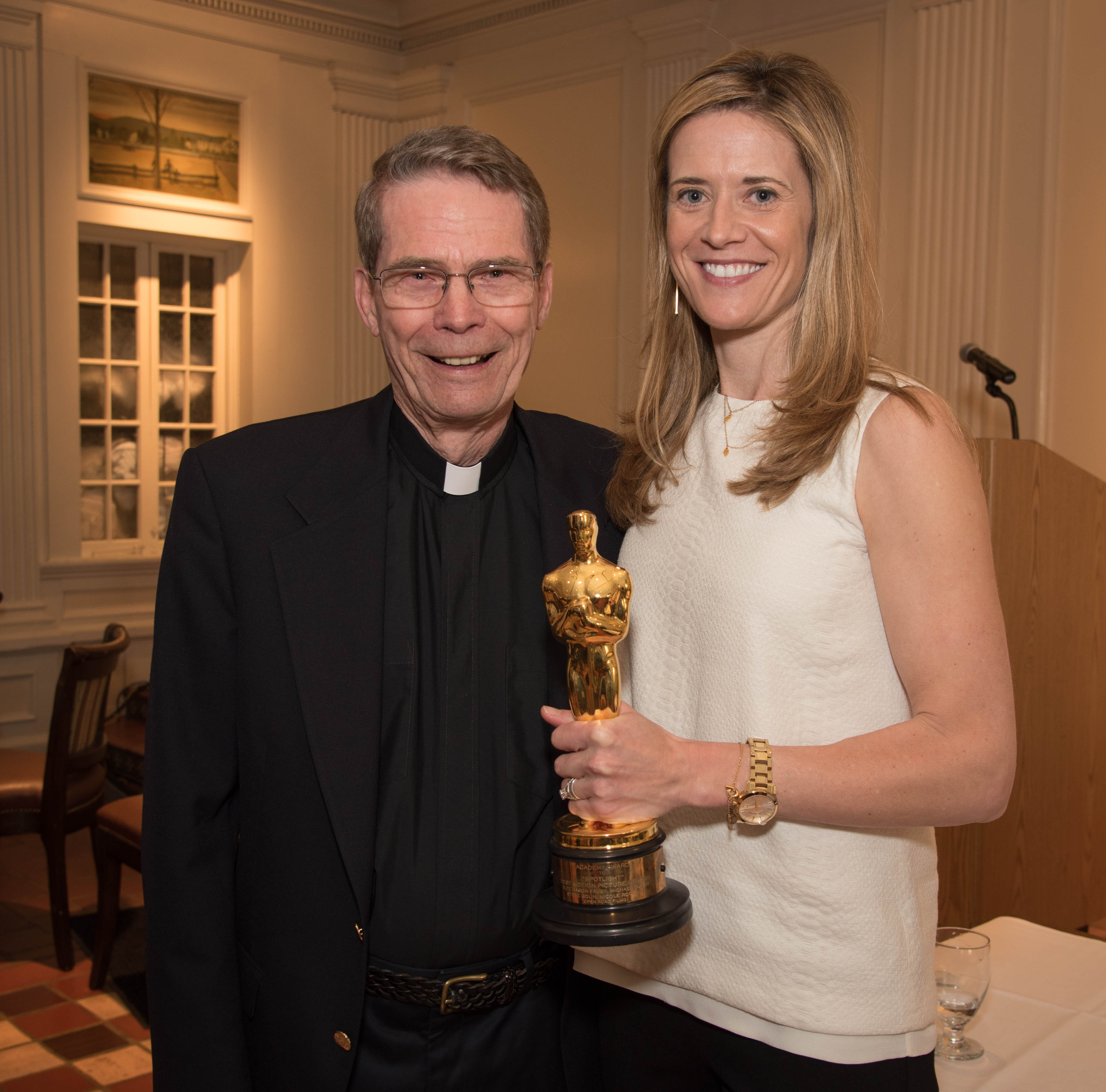 A woman holding a trophy stands next to a man in clerical attire.