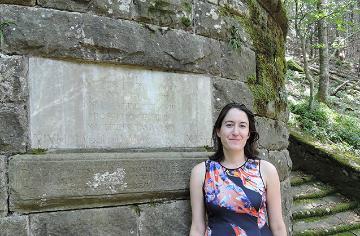 Woman standing beside ancient stone wall in a wooded area.