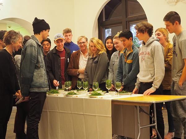 Table set with plates and wine glasses filled with grass with people surrounding it