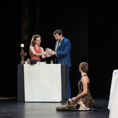 Four people performing a scene on stage with a table and props. image link to story