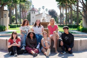 6 female and 1 male student playwrights from the SCU New Playwrights Festival sitting by the fountain in front of Mission Santa Clara Church