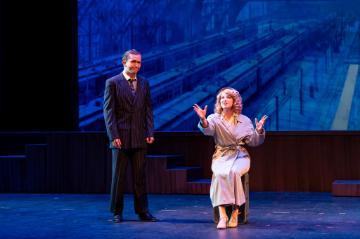 A man in a suit standing on the left and a woman in in a trench coat gesticulating and seated on the right against a bright blue background