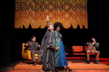2 men seated in armchairs on a stage with a black woman and a white woman standing