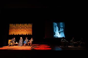 2 men and 2 women panelists seated in a spotlight on left of stage, with a black and white film showing above a darkened group of 3 people
