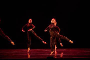 2 female dancers in black unitards lit in red on a black stage