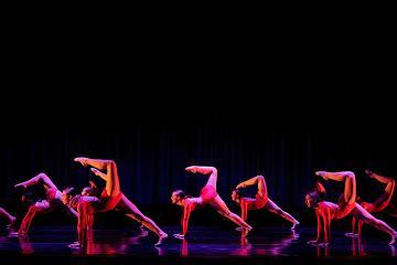 6 female dancers in red sparkly unitards with hands down & legs extended up like a scorpion tail