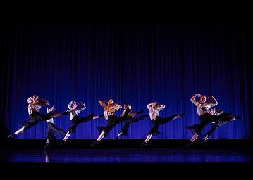8 dancers in white tops, colored scarves and black pants leaping diagonally across the stage