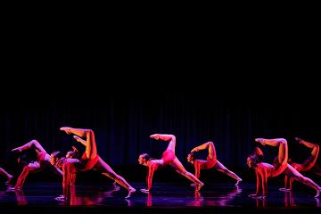 6 female dancers in red sparkly unitards with hands down & legs extended up like a scorpion tail
