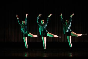 3 female dancers in bright green shirts and black shorts onstage with their hands lifted and