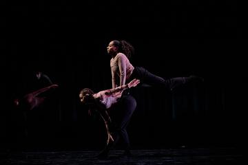 2 dance students in beige tops and black pants onstage, one jumping over the other