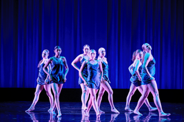 7 female dancers in short blue strappy dresses in a group onstage
