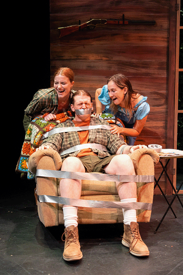 A man strapped with duct tape in a recliner with 2 women laughing looking on