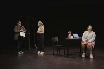 Ina black box theatre, one woman entered through a door to meet a second woman who's standing and a third who's seated in the bcakground