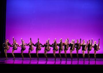 A kick line of 14 female dancers in black sparkly dresses, black fishnets and heeled tap shoes