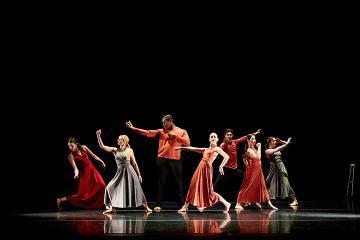 A group of 5 female dancers in long dresses and 2 men in red shirts and black pants onstage