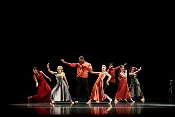 A group of 5 female dancers in long dresses and 2 men in red shirts and black pants onstage