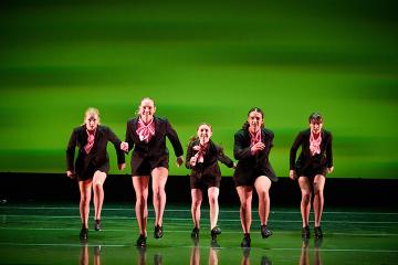5 tap dancers in black sequined costumes dancing onstage with a bright green background