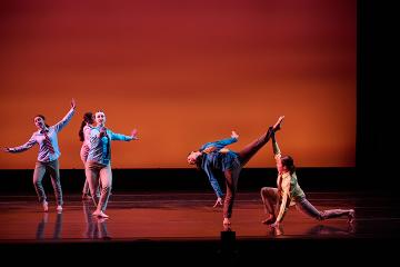 4 female modern dancers in light shirts and dark pants spread across a stage with arms extended