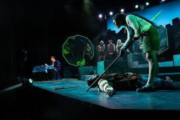 A woman with a lance standing over a fallen man with a Dungeon Master and D&D paraphernalia in the background