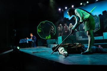 A woman with a lance standing over a fallen man with a Dungeon Master and D&D paraphernalia in the background
