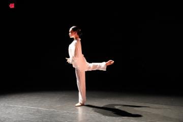 Femal dancer in white looking down at her hands with one leg bent and lifted behind her