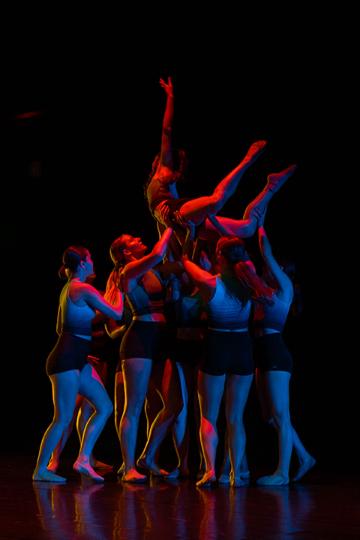 A female dancer onstage being held aloft by 6 others, all wearing white tops and black shorts, lit in red