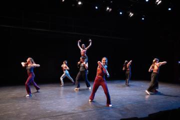 A group of 6 female modern dancers in bright unitards dancing in a black box theatre