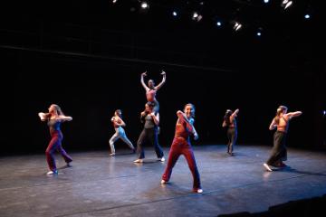 A group of 6 female modern dancers in bright unitards dancing in a black box theatre