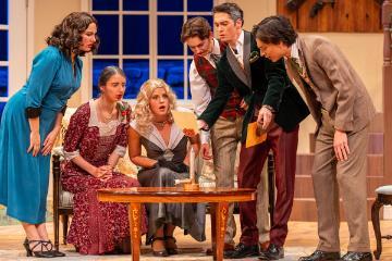 3 men and 3 women in 1930s costumes peering over a table with a ouija board on it
