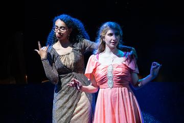 One woman in a gold and black wraparound dress and another in a coral prom dress
