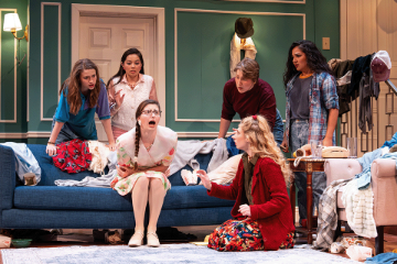 5 women surrounding a blue couch looking at a seated woman in a messy room with clothes draped everyehere
