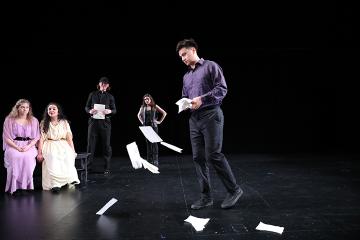 A man in a purple shirt and grey pants throwing up a sheaf of papers watched by 2 women seated and a man and a woman standing behind
