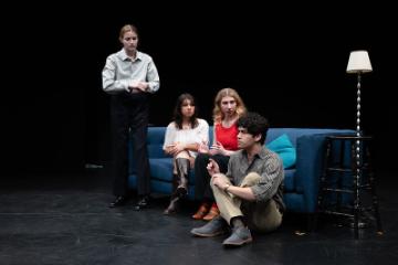 2 women sit on a blue counch and a third womane stands next to it, all looking at a man seated next to the couch on the floor