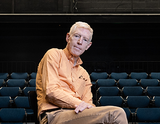 Brian Thorstenson sitting in Fess Parker studio Theatre wearing an orange shirt, khaki pants and Converse boots image link to story