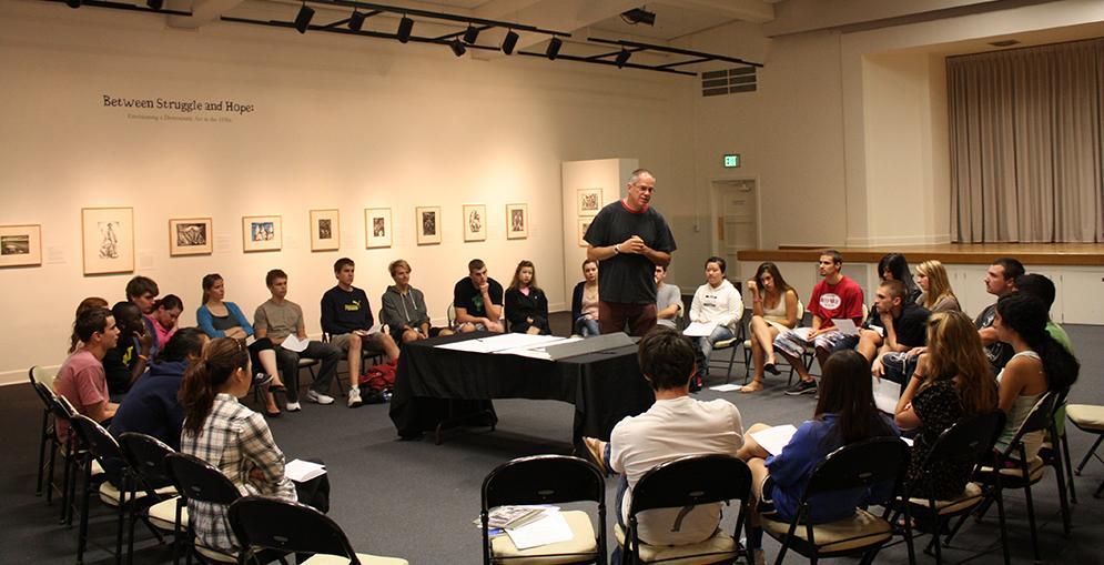 Students sitting in a circle, listening to guest curator Art Hazelwood talk about an exhibition.