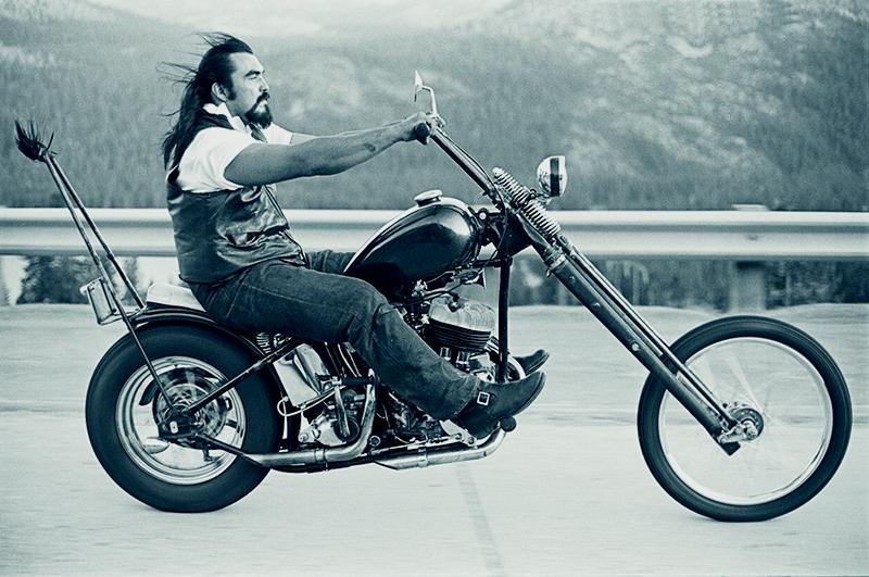 Black and white photograph of man on motorcyle on freeway with mountains in background.